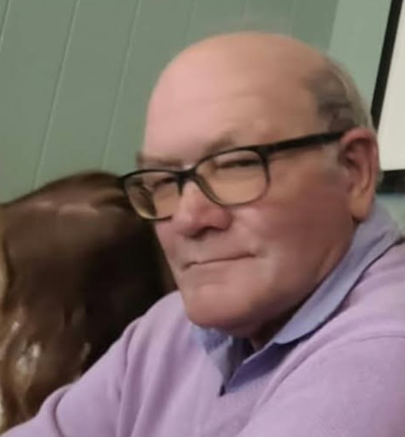 Portrait photo of John Robbins, smiling, wearing glasses and a lilac jumper